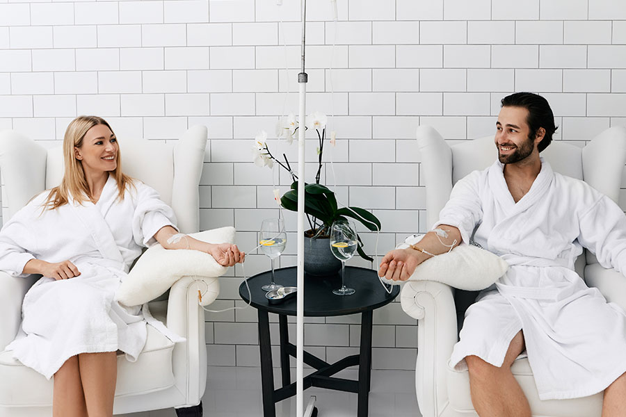 A young couple sits in separate white chairs with their arms extended and smiling as they receive vitamin IV infusion therapy at a modern wellness center in Fort Worth, TX.