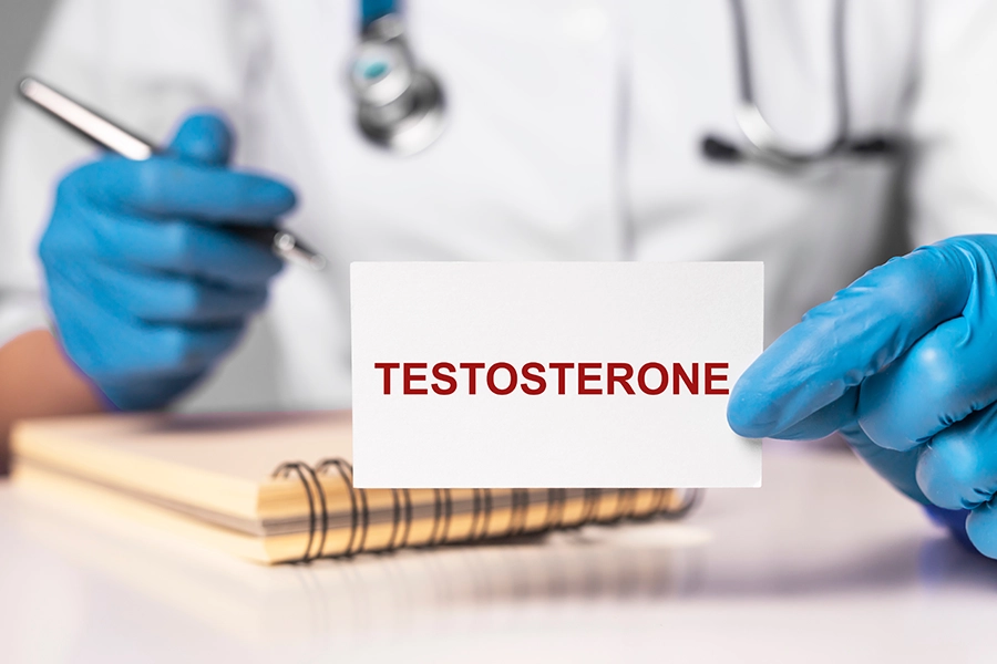 A doctor's gloved hands hold a pen and a card that says "testosterone" in an office in Farmer's Branch, TX.