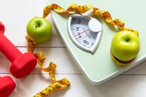 Simply Direct Medicine – a scale on the ground with two green apples and measuring tape sitting on top of it and red weights next to it, symbolizing weight loss.